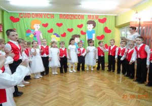Dziewczynki w białych sukienkach oraz chłopcy w czerwono-czarnych kamizelkach i czarnych spodniach w półkolu recytuja wiersz dla rodziców.